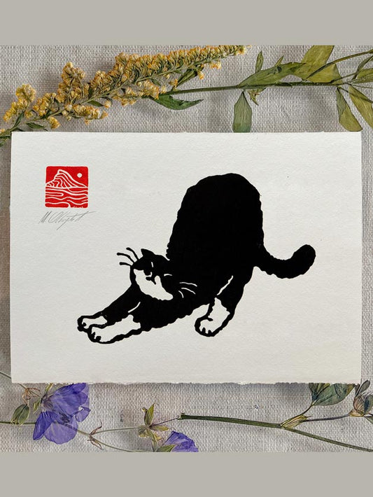 A square white art print featuring a black cat silhouette stretching, with a red square mountain-style logo in the upper left. The print is styled on a textured fabric background with dried yellow flowers and green leaves around it.