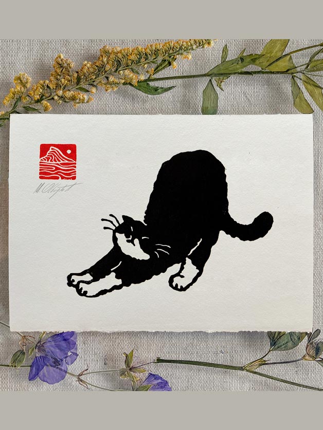 A square white art print featuring a black cat silhouette stretching, with a red square mountain-style logo in the upper left. The print is styled on a textured fabric background with dried yellow flowers and green leaves around it.
