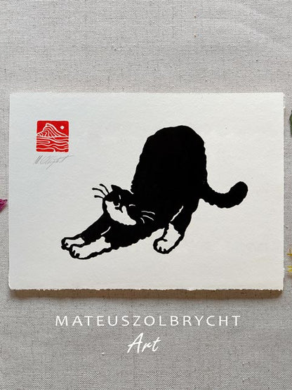 A square art print featuring a black cat silhouette stretching on an off-white background, with a small red square stamp in the upper left. The print is photographed on a textured light gray fabric surface, with the text 'MATEUSZOLBRYCHT Art' printed below.